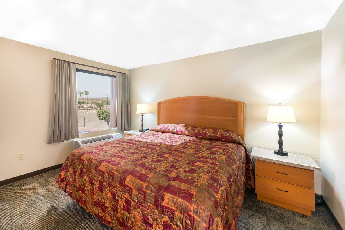 A hotel room with a large bed, two bedside tables with lamps, a window with curtains, and a view of palm trees.