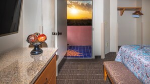 Habitación de hotel con cama, un banco, un escritorio con un globo terráqueo y vistas a un atardecer a través de una puerta abierta.