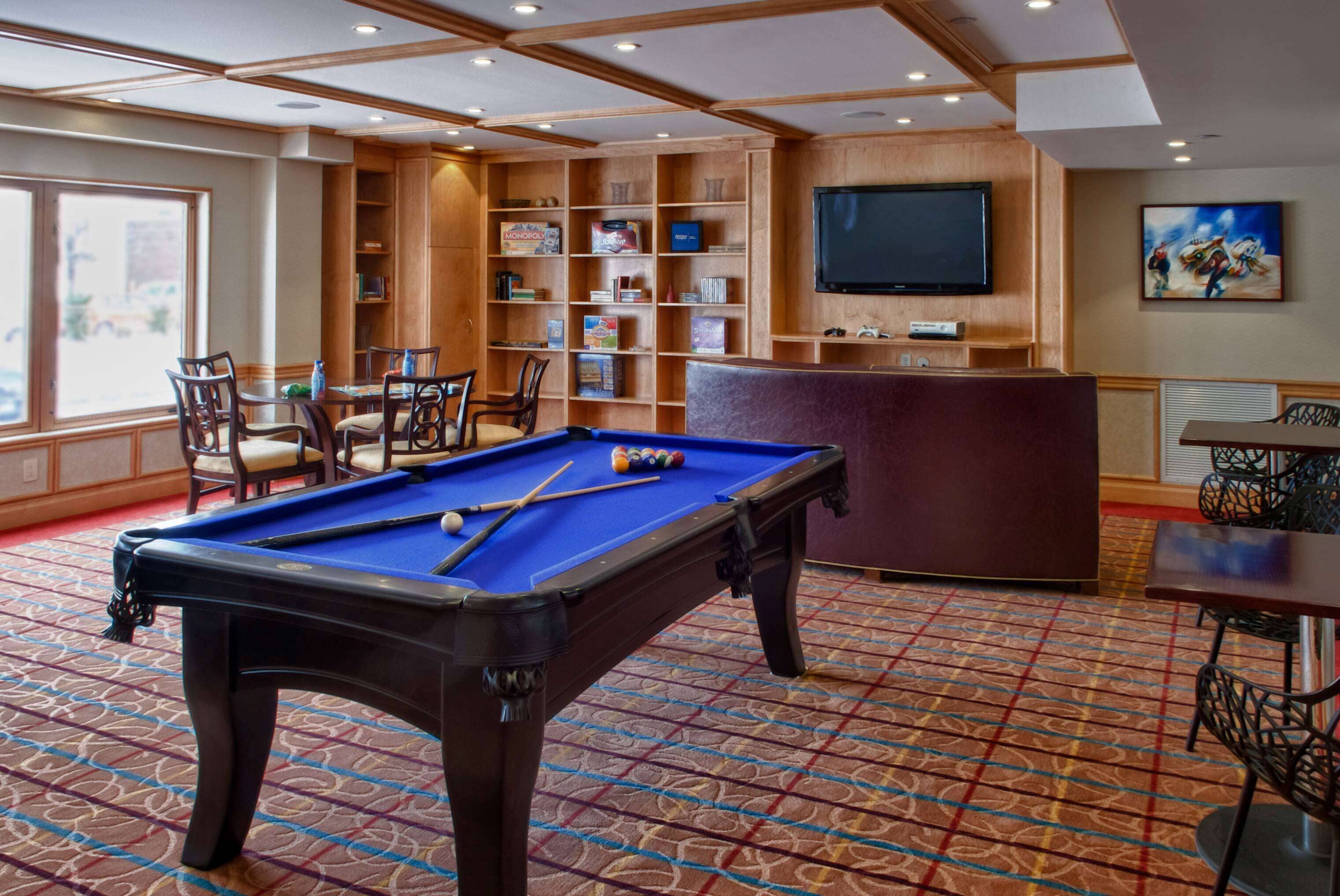 A pool table with chairs, a bookshelf, a television, and a painting in a room with patterned carpet.