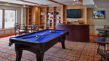 A pool table with chairs, a bookshelf, a television, and a painting in a room with patterned carpet.