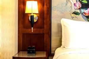 Una mesita de noche de madera con una lámpara y un pequeño objeto negro encima, una cama blanca con un cojín blanco, y una pared con una obra de arte floral.