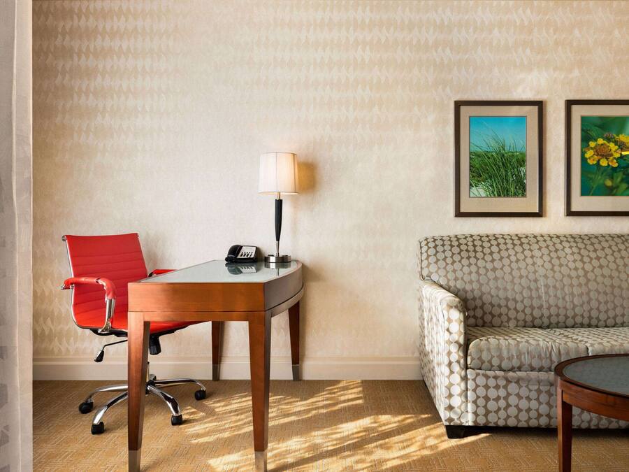 A hotel room with a red office chair, a wooden desk with a lamp, a patterned sofa, and two framed pictures on the wall.