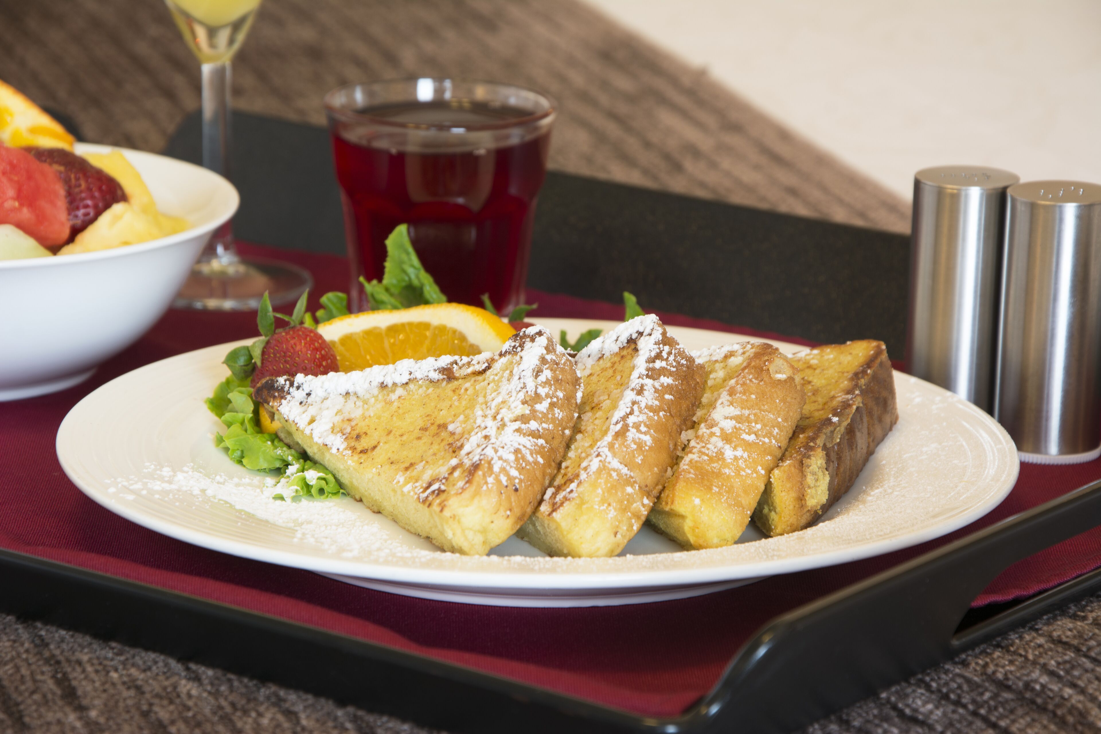 A plate of French toast with powdered sugar, accompanied by a bowl of fruit and a glass of red beverage, on a table with a red napkin and salt and pepper shakers.