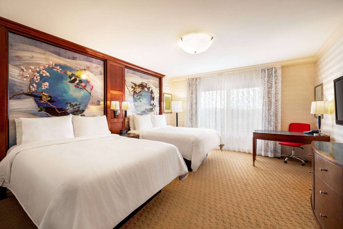 Een hotelkamer met twee bedden, een bureau, een stoel, een televisie en kunstwerken aan de muur.