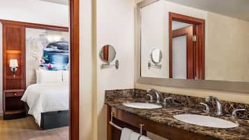 A hotel bathroom with two sinks, a large mirror, and a bed with a nightstand.