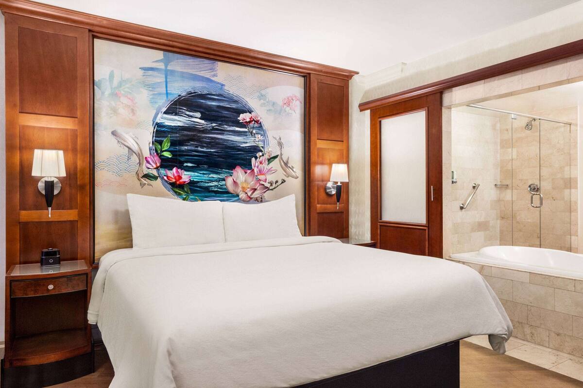Hotelkamer met een groot bed, een schilderij van water en bloemen aan de muur, een badkamer met bad en douche, en houten lambrisering.