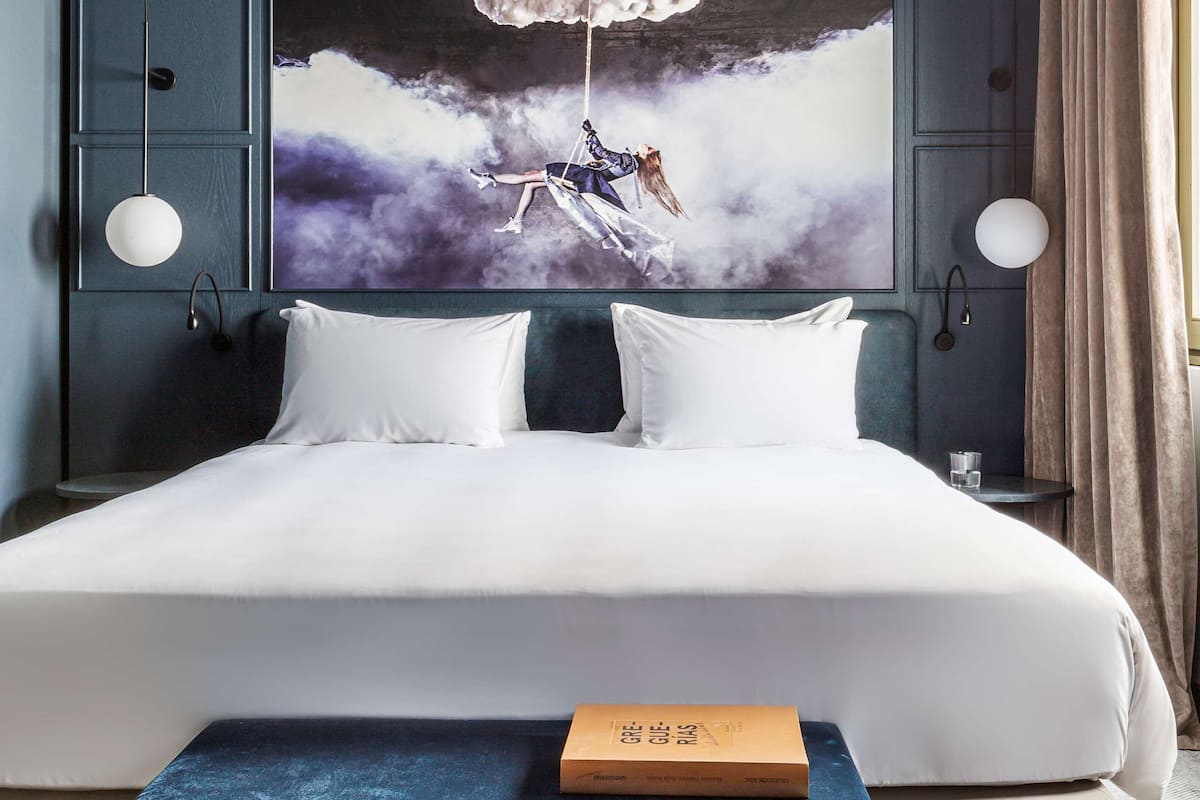 Una camera d'albergo con un letto grande, un comodino con un libro, un quadro appeso al muro e un tavolino da notte con una lampada.