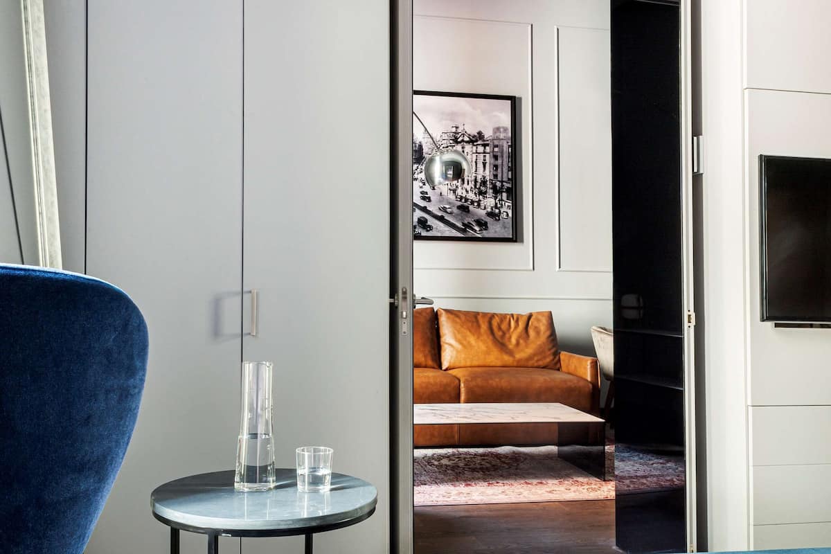 Una camera d'albergo con un letto, una poltrona blu, un tavolino rotondo con un bicchiere e un quadro appeso alla parete.