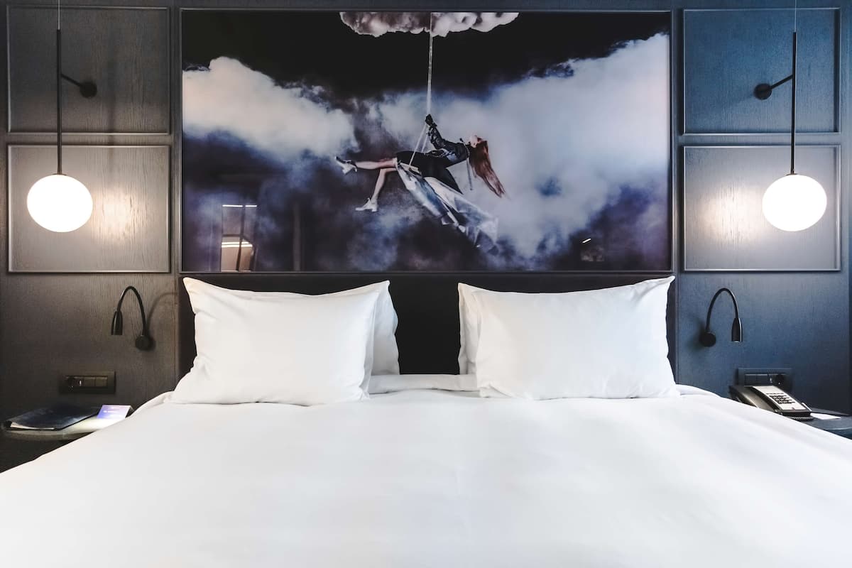 Una camera d'hotel con un letto grande, biancheria bianca, un quadro a parete raffigurante una persona su un'altalena, due abat-jour e un telefono sul comodino.