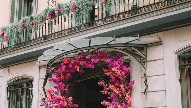Entrada de un edificio con balcón de hierro forjado, un letrero azul con 'H' y 'BLU', y plantas florecientes de color rosa.