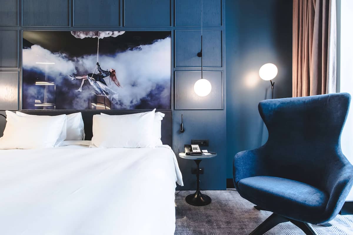 Una camera d'albergo con un letto, una poltrona blu, un tavolino con un telefono e un lampadario a parete.