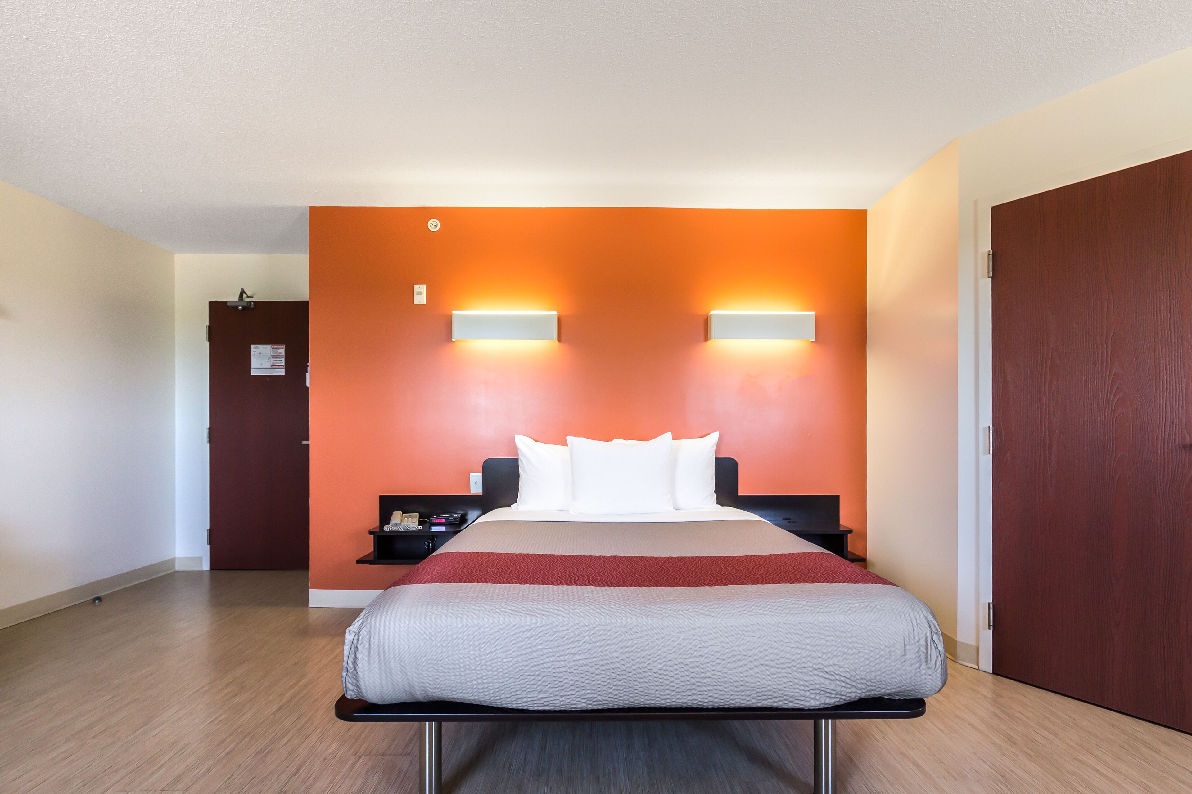 A hotel room with a bed, a nightstand, a wall-mounted light, and a door.