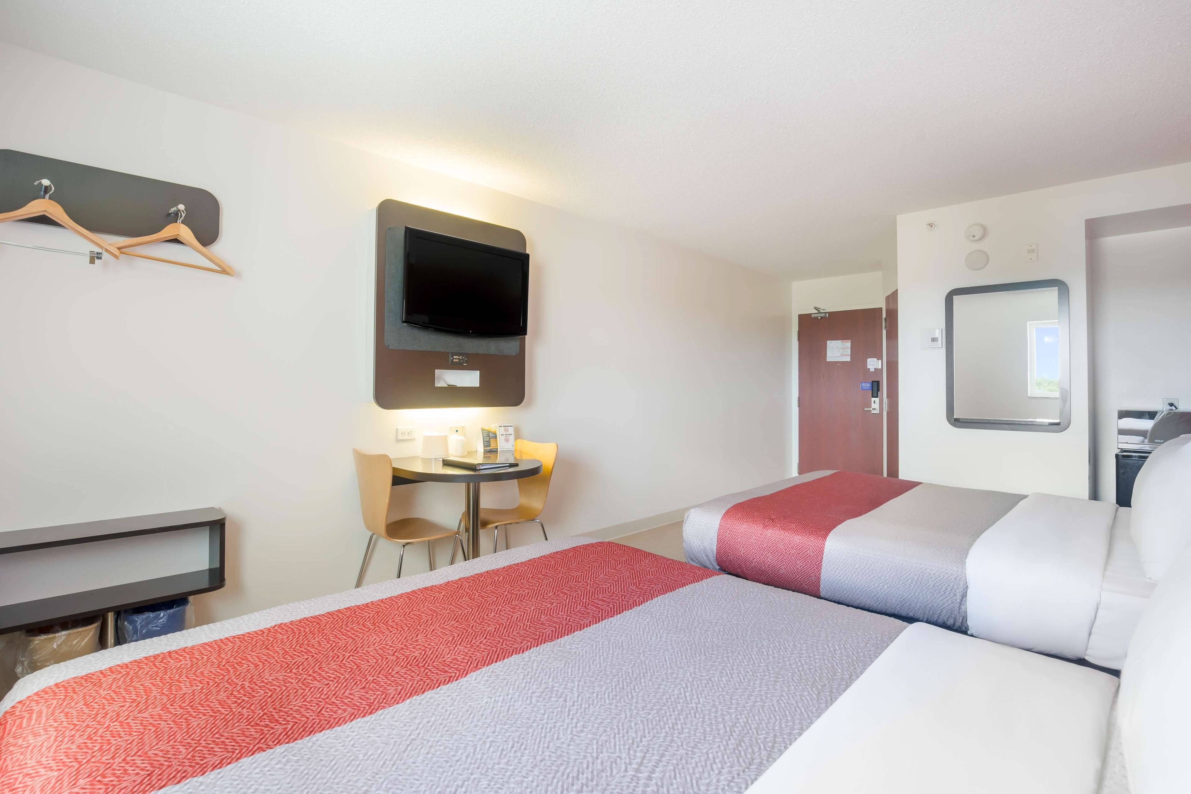 A hotel room with two beds, a TV mounted on the wall, a desk with chairs, and a small table with a lamp.