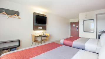 A hotel room with two beds, a TV mounted on the wall, a desk with chairs, and a small table with a lamp.