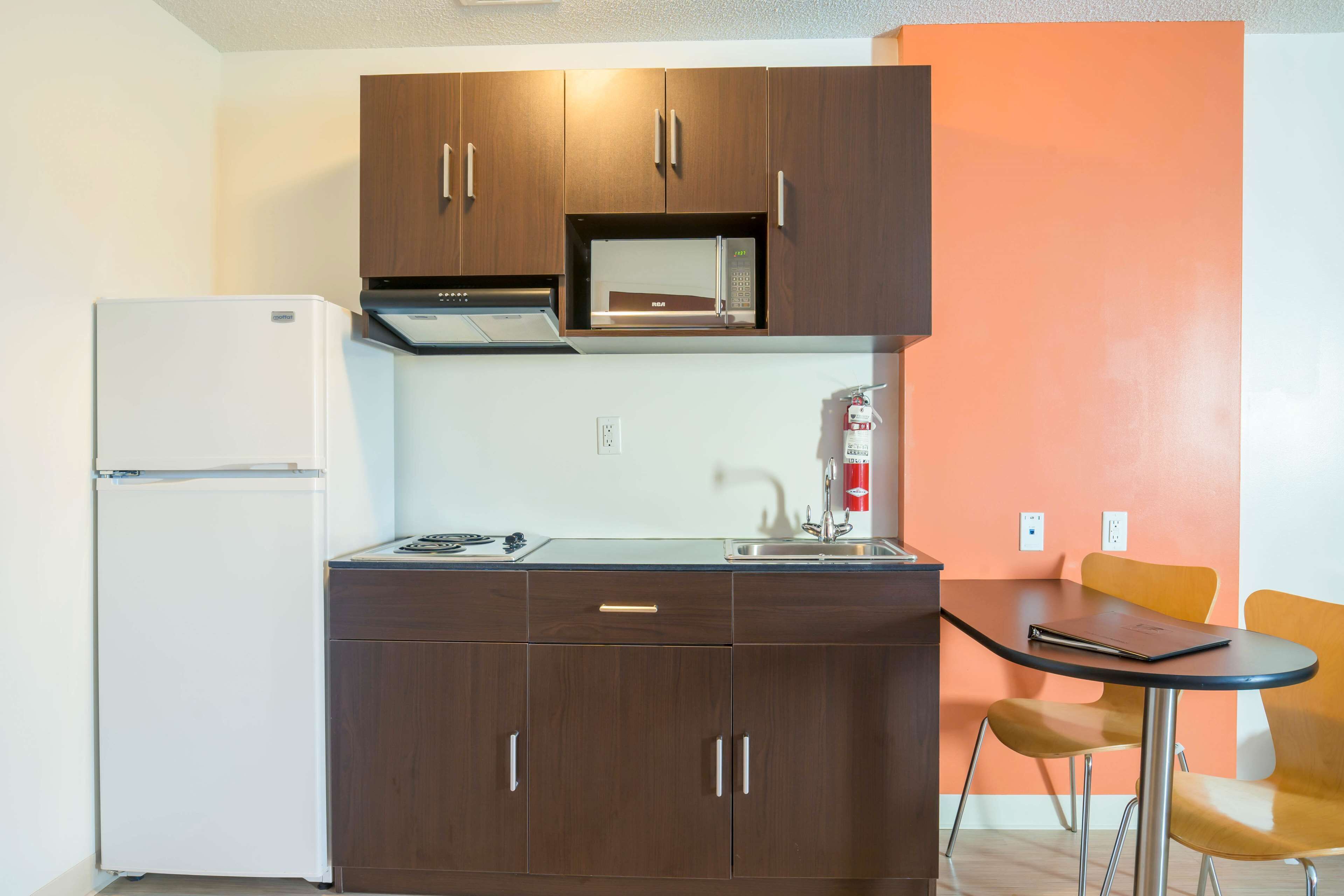 A compact kitchen with a refrigerator, microwave, and sink. There is a small table with chairs and a fire extinguisher on the wall.