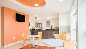 Un comedor moderno con paredes naranjas, mesas blancas y sillas amarillas. Un televisor de pantalla plana está montado en la pared.