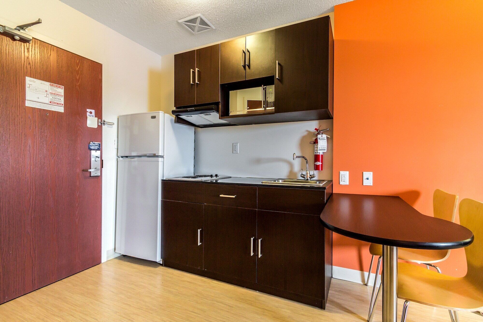 Una cocina compacta con refrigerador, microondas y lavamanos. Hay una pequeña mesa de comedor con dos sillas.