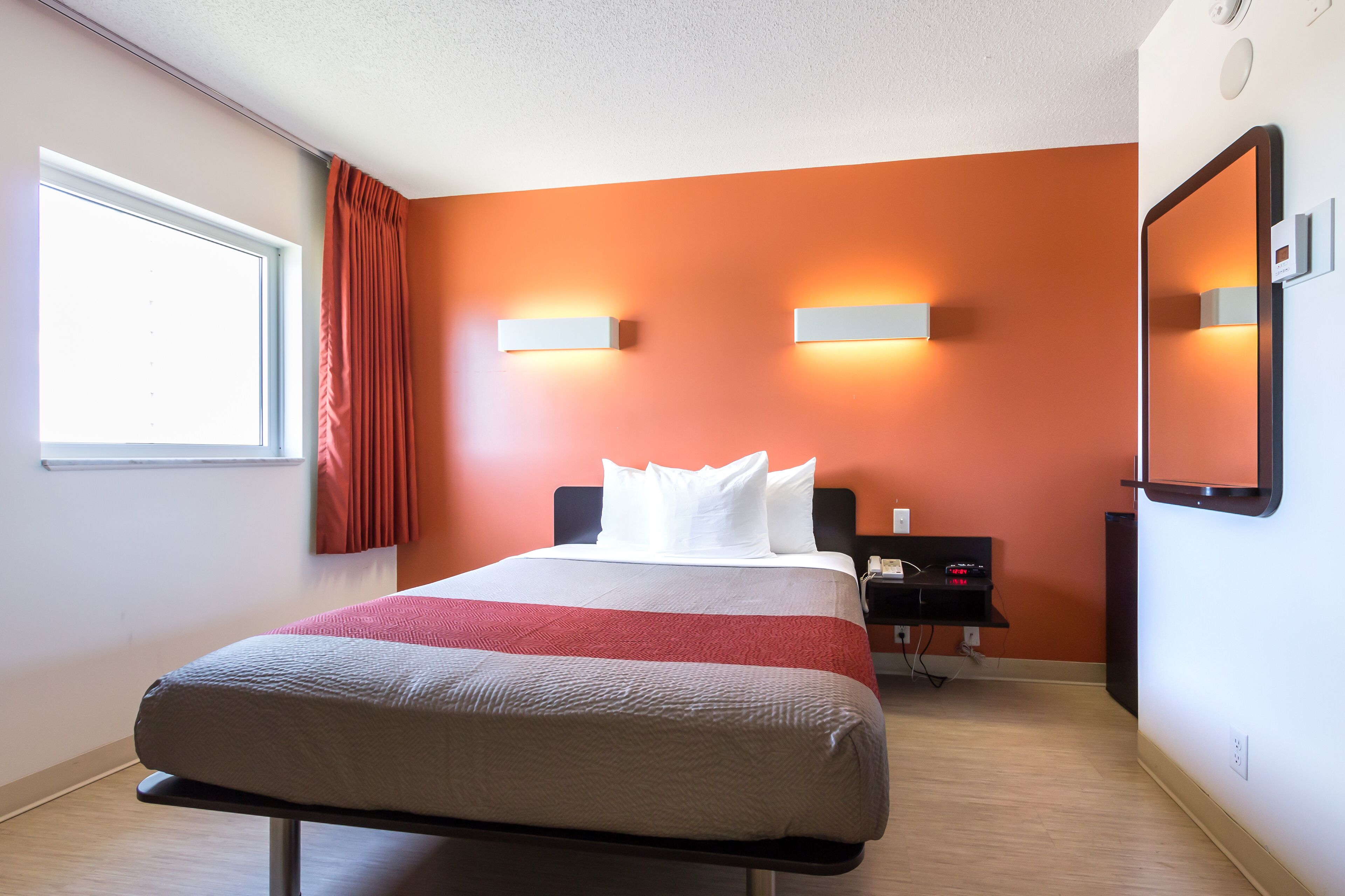 A hotel room with a bed, a nightstand, a wall-mounted mirror, and a window with curtains.