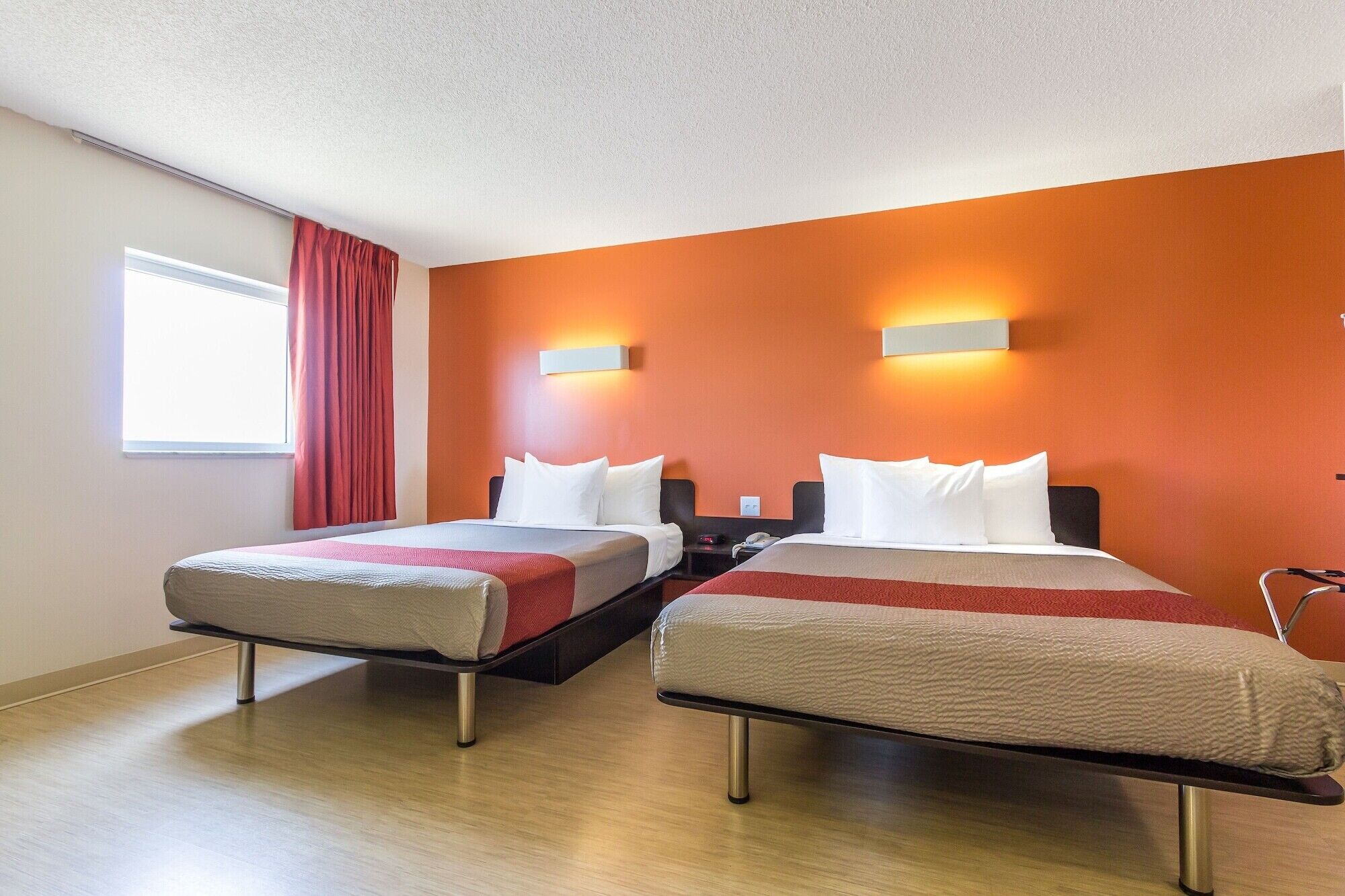 A hotel room with two beds, a window with red curtains, and an orange accent wall.