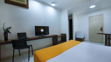 Habitación de hotel con cama, escritorio, ordenador portátil, televisor, silla, planta en maceta y puerta.