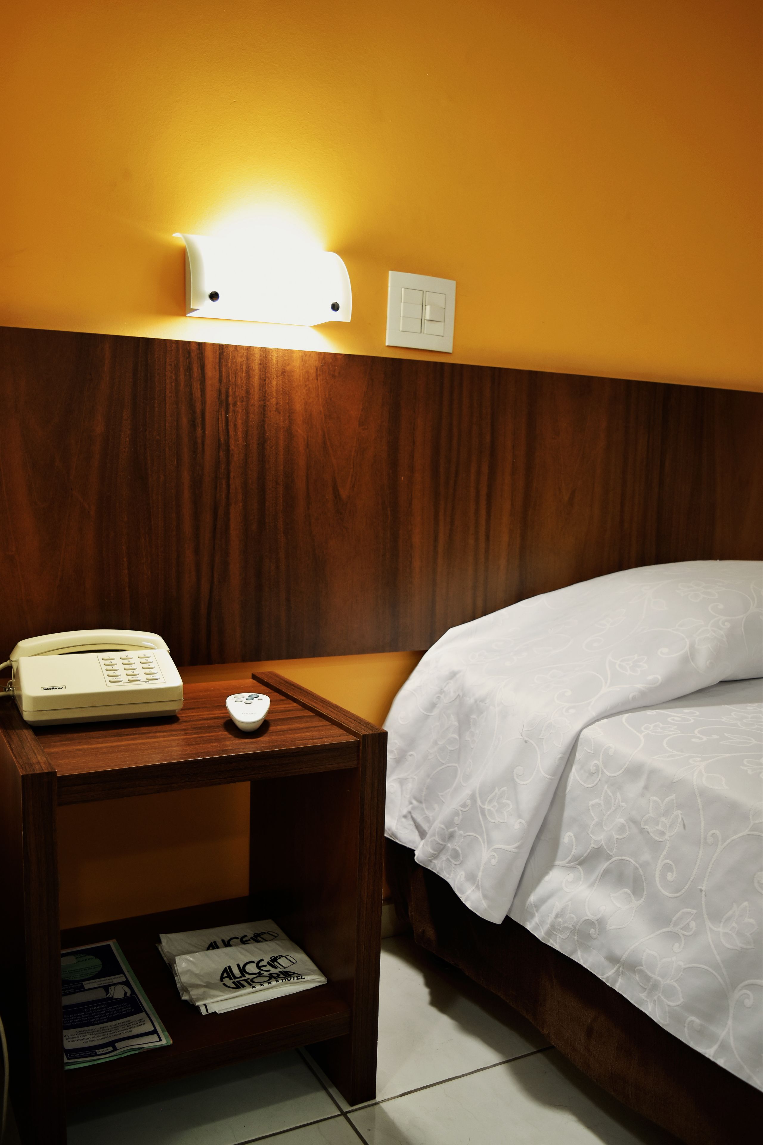 A hotel room with a wooden headboard, a bedside table with a telephone and a remote control, and a bed with a white bedspread.