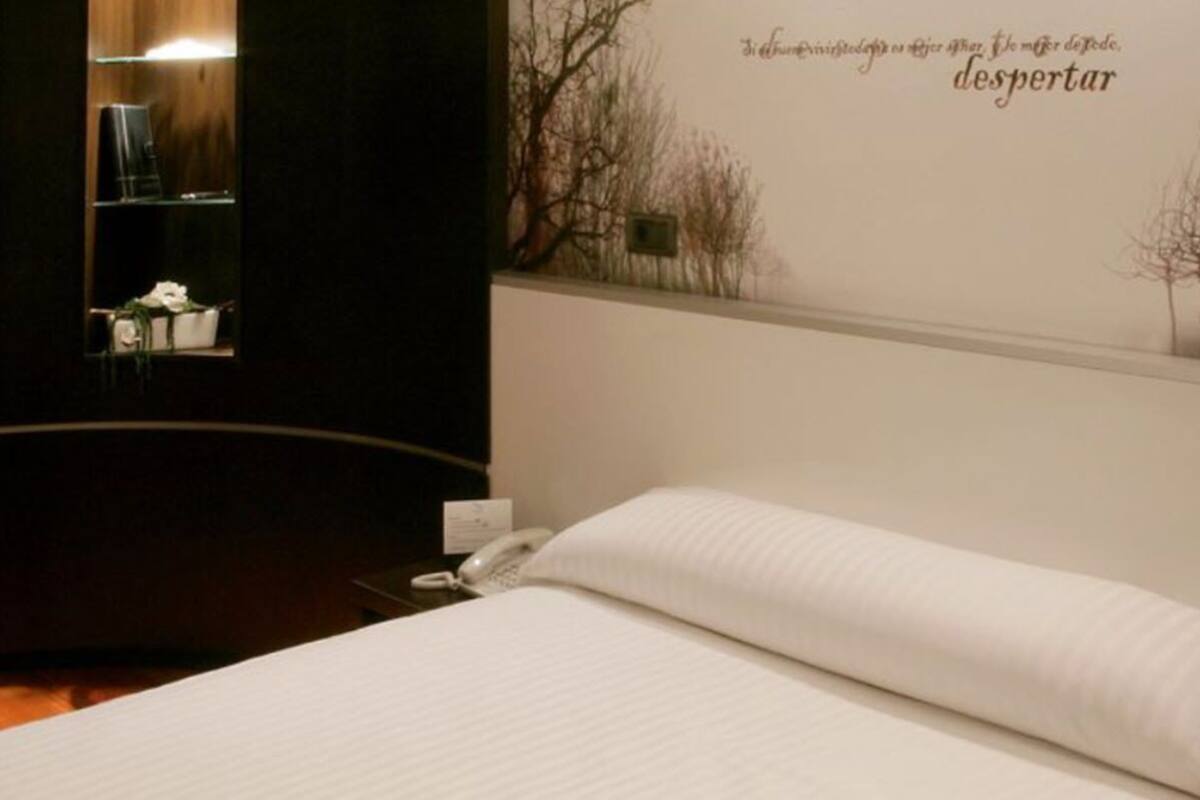A hotel room with a bed, a desk with a chair, a small table with a plant, and a wall with a tree and text.