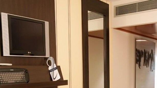 A modern hotel room with a large mirror, a flat-screen TV, a desk with a keyboard, and a bench.