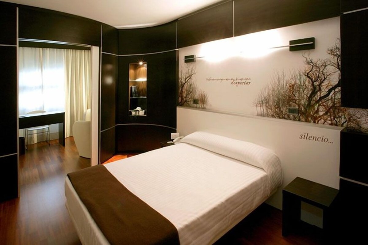 A modern bedroom with a large bed, a desk, and a chair. There is a wall with a decorative light and a sign that reads "silencio."