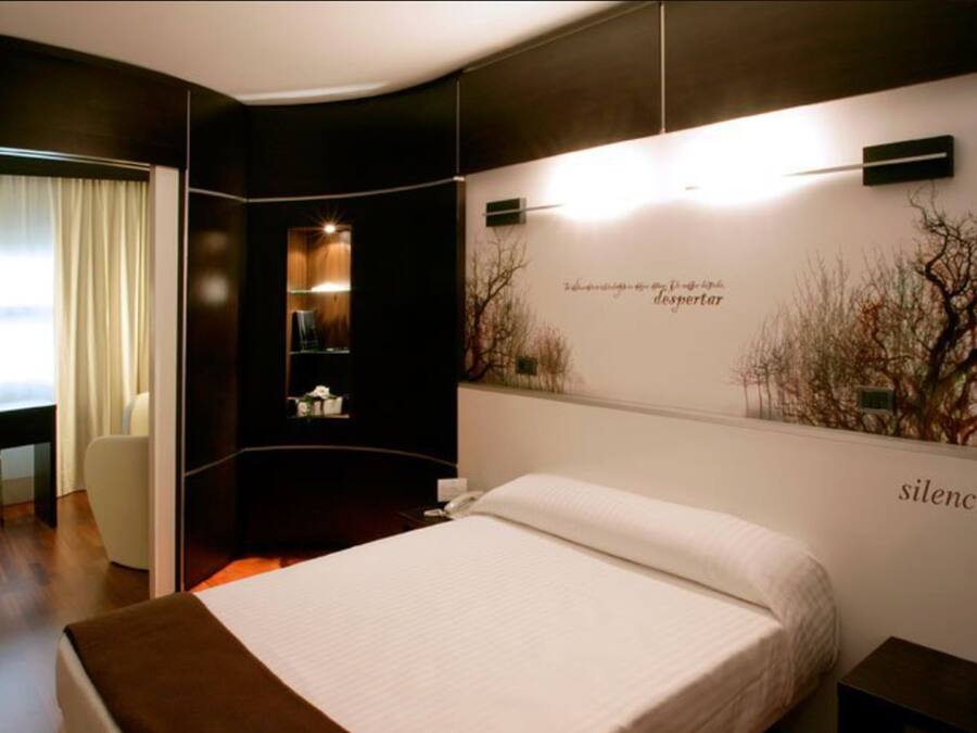 Ein modernes Hotelzimmer mit einem großen Bett, einem Schreibtisch und einem Stuhl. Eine Wand mit dekorativer Beleuchtung und einem Schild, auf dem 'silencio...' steht.