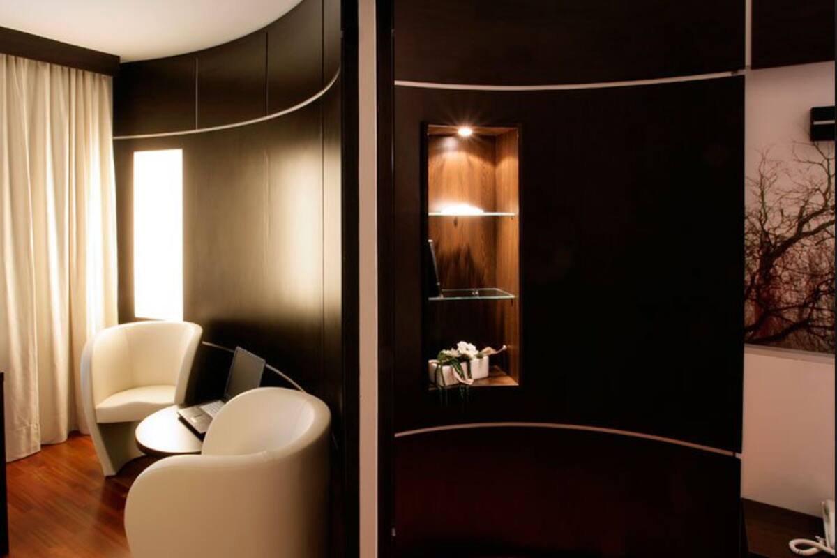 A modern room with a curved wall, a white armchair, a small round table with a laptop, and a window with curtains.