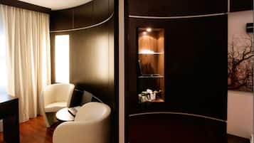 Una habitación moderna con una pared curva, un sillón blanco, una pequeña mesa redonda con una computadora portátil y una ventana con cortinas.