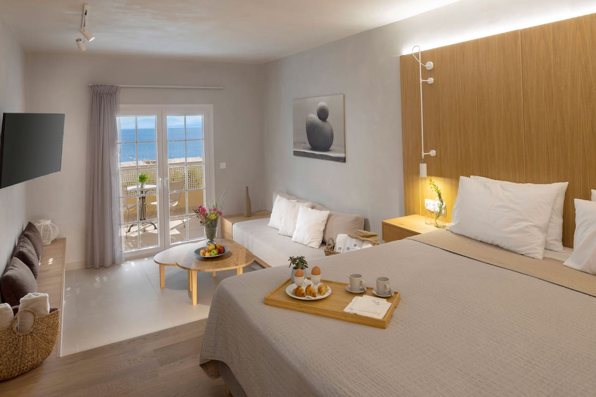 A hotel room with a bed, a bedside table, a TV, a balcony with a view of the sea, and a breakfast tray on the bed.
