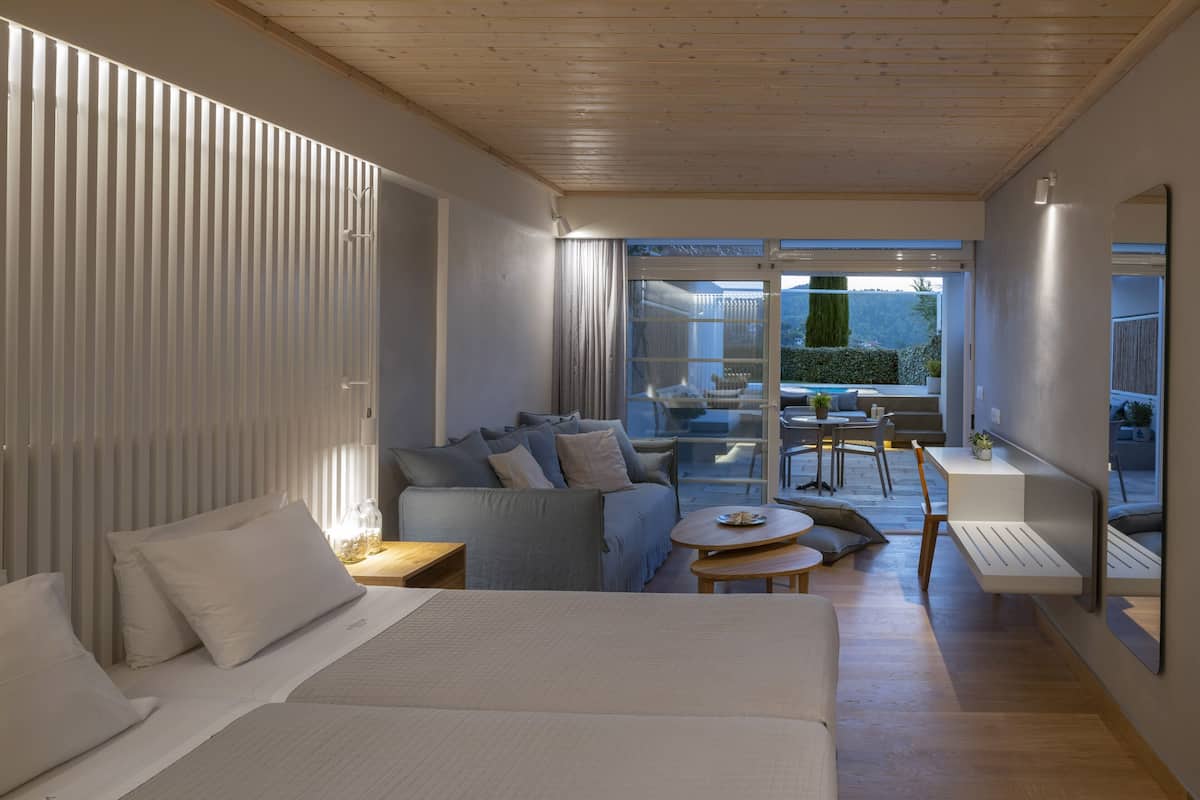 A modern hotel room with a large bed, a sofa, a dining area with a table and chairs, and a balcony with a view of the outdoors.