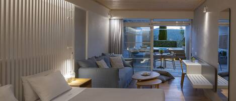A modern hotel room with a large bed, a sofa, a dining area with a table and chairs, and a balcony with a view of the outdoors.