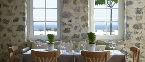 A dining table set with white tablecloth, chairs, and glassware, with two windows offering a view of the sea.