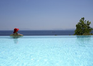 A person in a red hat relaxing in a clear blue infinity pool with a view of the ocean and a tree.