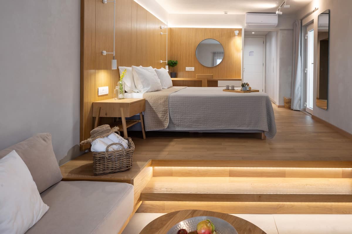 A modern hotel room with a wooden floor, a bed with a grey bedspread, a wooden desk, a wicker basket, and a round table with fruits.