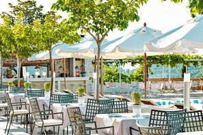 An outdoor dining area with tables and chairs, umbrellas, and a pool in the background.