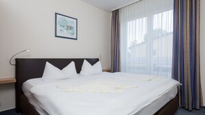 A neatly made bed with white linens and a checkered blanket, a headboard, a bedside table, and a window with curtains.