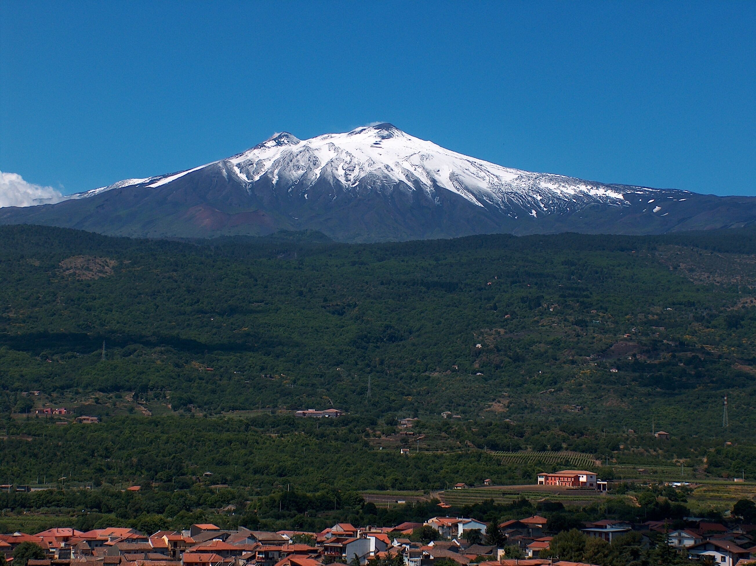 Une montagne aux sommets enneigés, surplombant un ciel dégagé, entourée d’une zone densément boisée et d’un petit village aux maisons aux toits rouges.