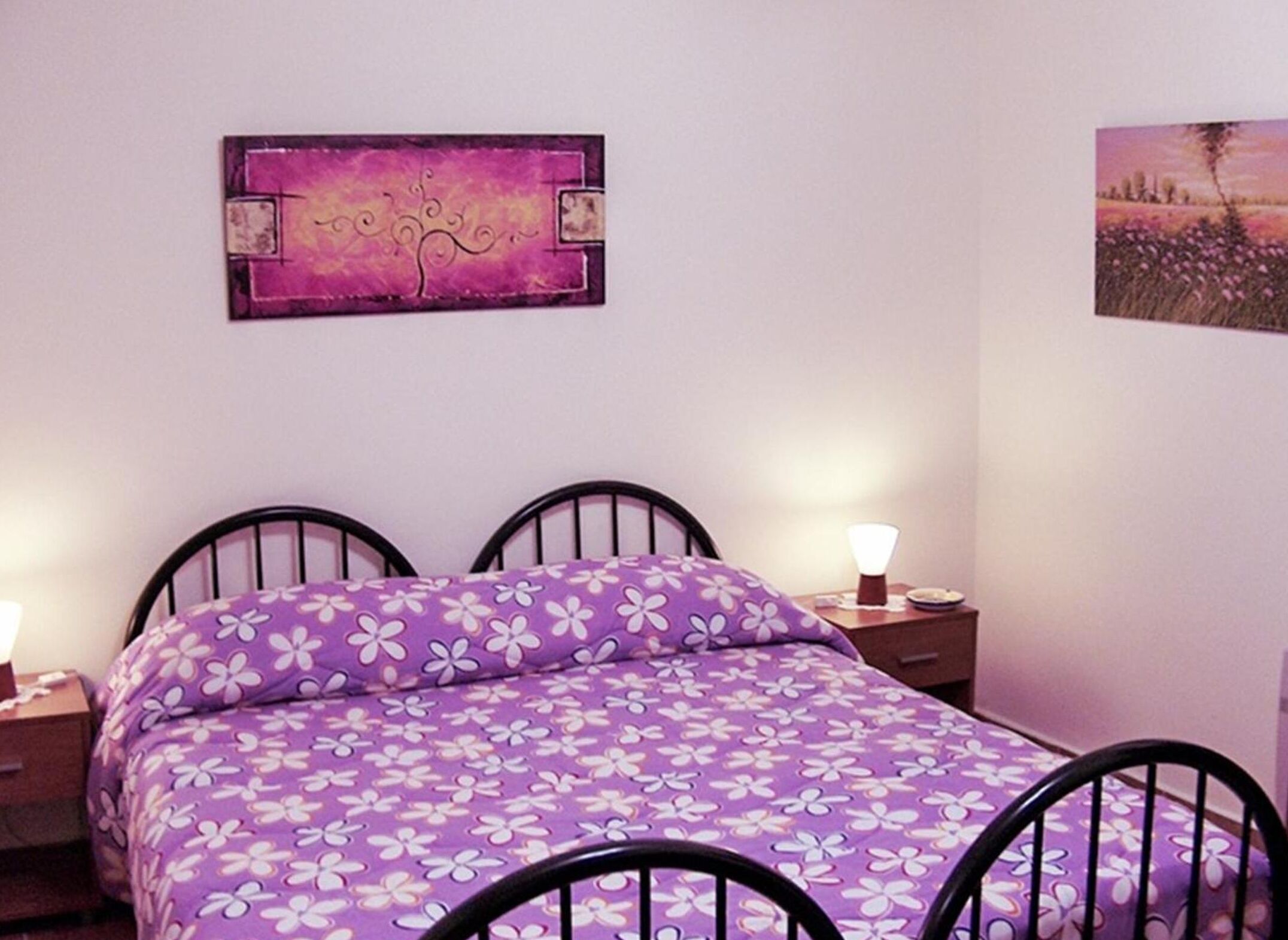 Ein Schlafzimmer mit violett geblümtem Bettbezug, zwei schwarzen Metall-Kopfteilen und Nachttischlampen.