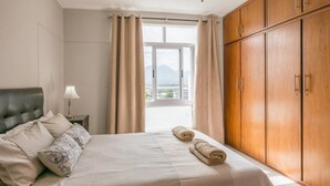 A bedroom with a bed, a nightstand, a lamp, a window with curtains, and a wardrobe.