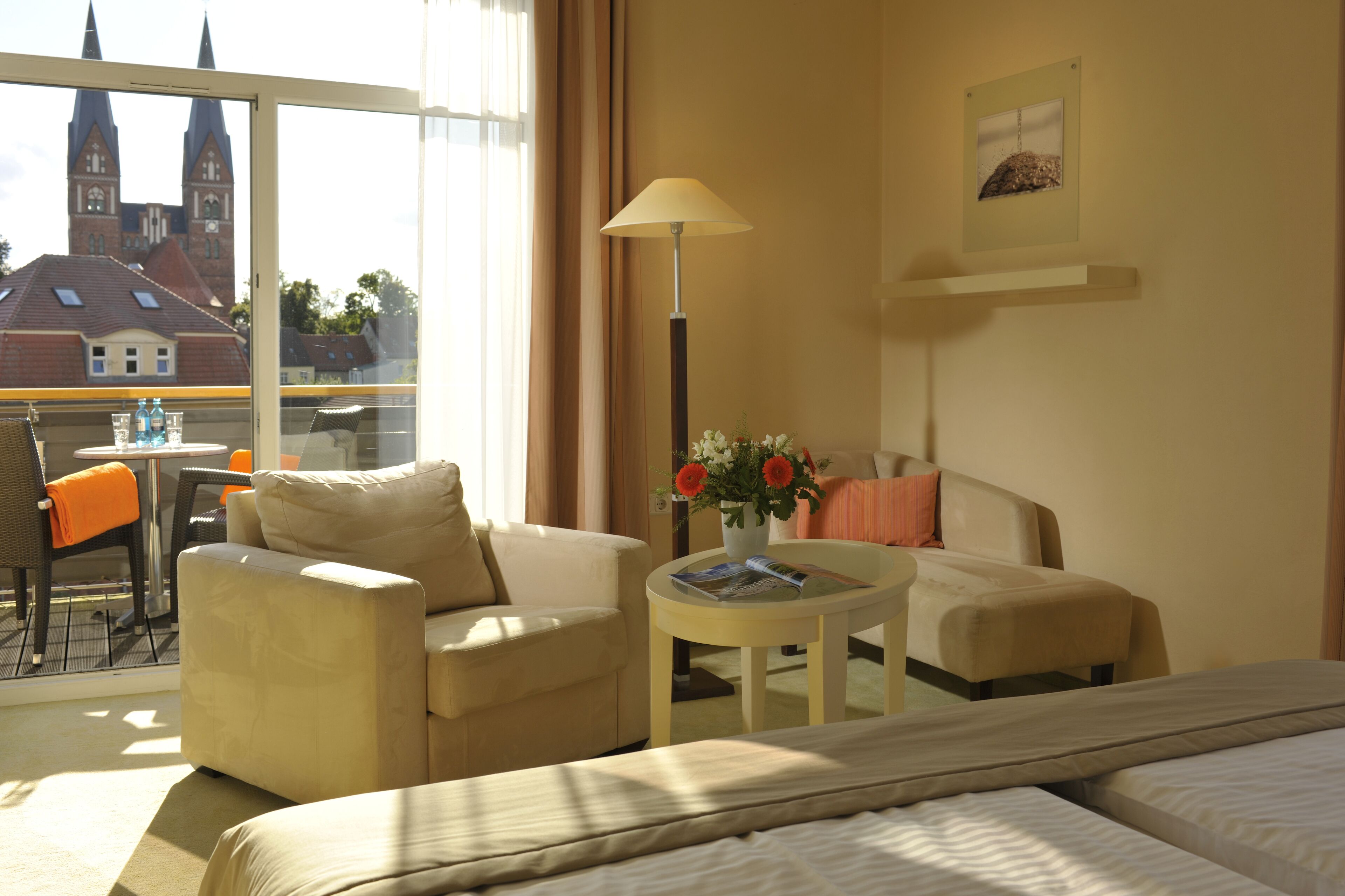 A hotel room with a bed, two armchairs, a small table with a vase of flowers, and a view of a church and buildings outside.