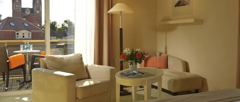 A hotel room with a bed, two armchairs, a small table with a vase of flowers, and a view of a church and buildings outside.