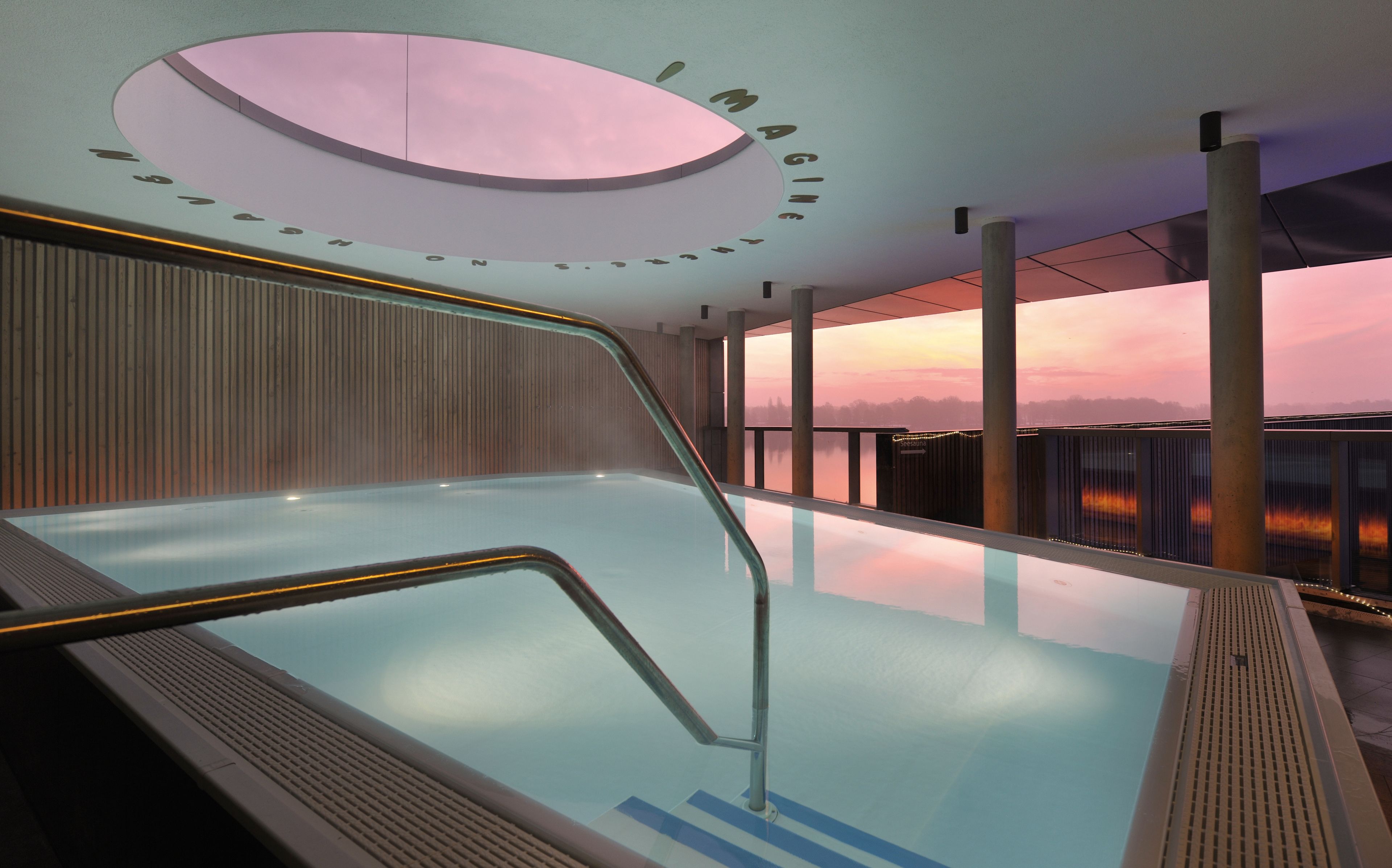 Uma piscina interior moderna com barreira de vidro, parede de madeira e vista para o pôr do sol.