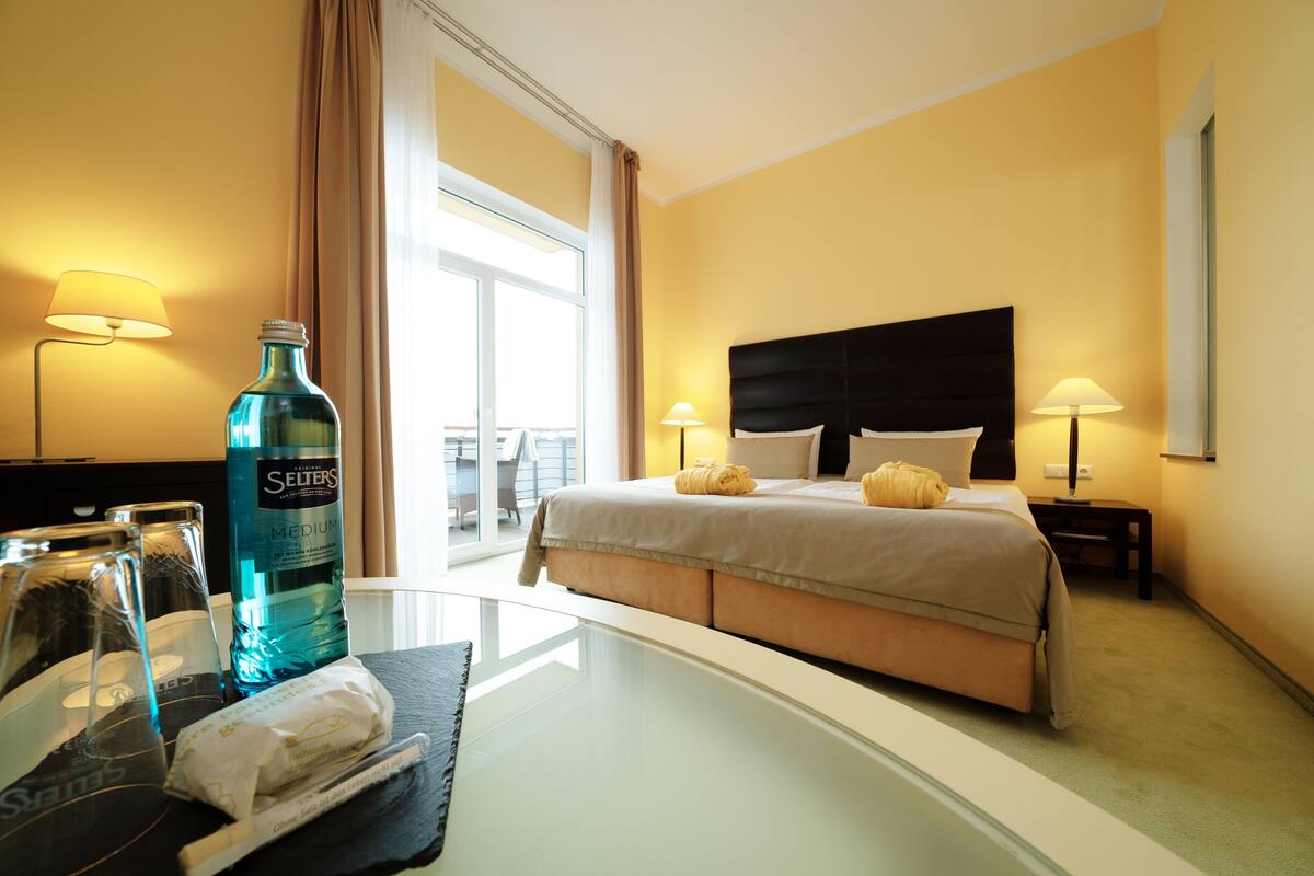 Ein Hotelzimmer mit einem Bett, zwei Nachttischlampen, einem Glastisch mit Flasche und Gläsern sowie Blick ins Freie.