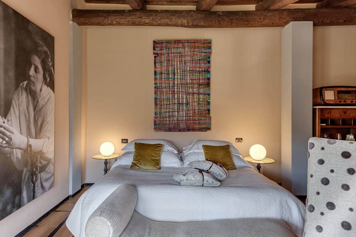 A bedroom with a bed, two bedside lamps, a large abstract painting, and a vintage radio.