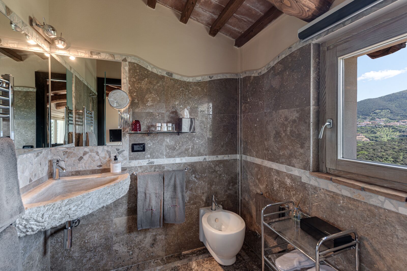 A bathroom with a stone sink, a bidet, a mirror, and a window with a view of the countryside.