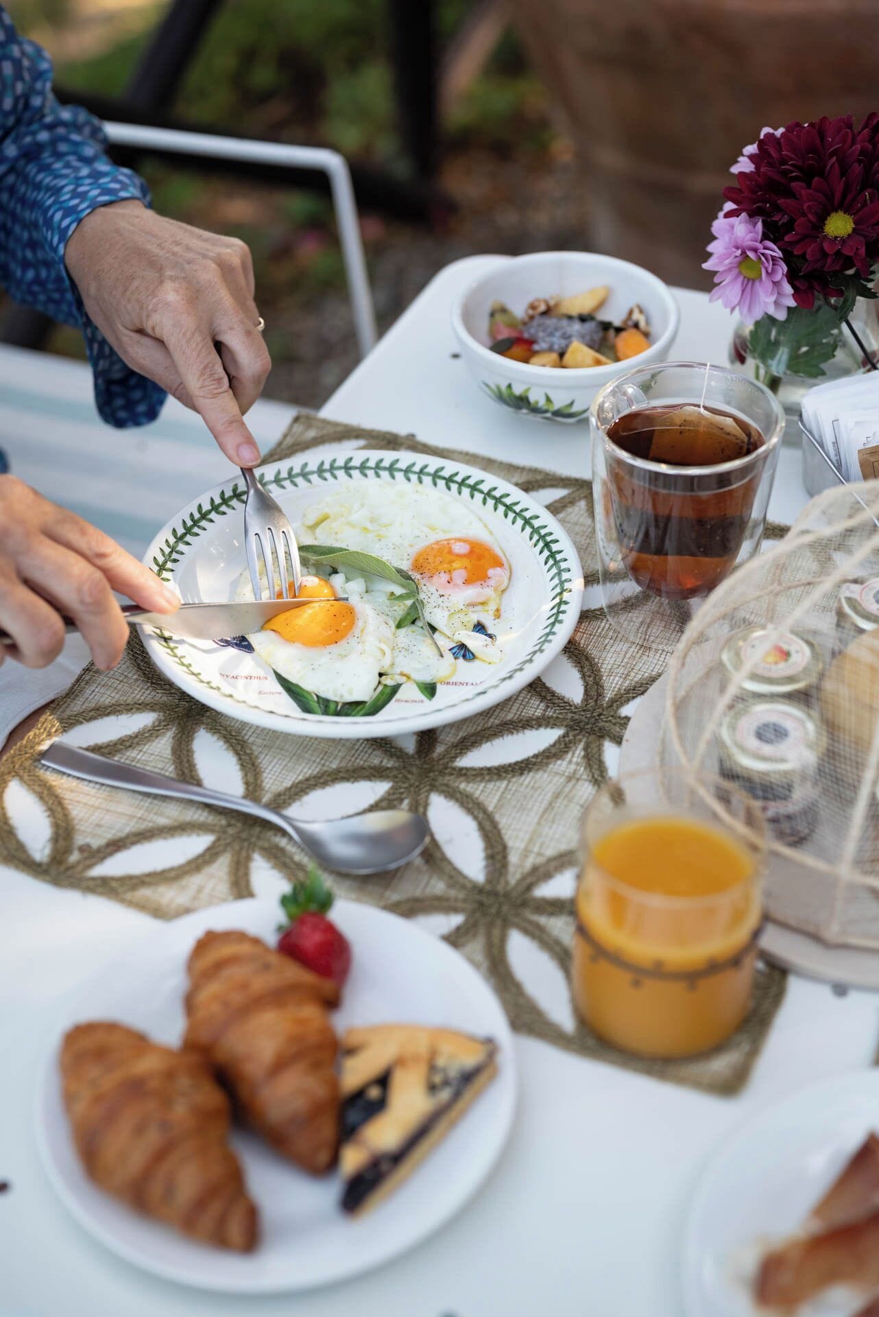 A breakfast setting with a plate of eggs, a cup of tea, croissants, and a bowl of fruit.
