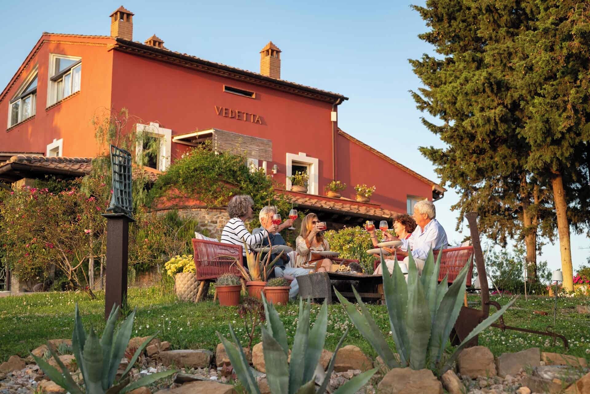 A red building with the name "VEDETTA" on the facade, surrounded by a garden with various plants and a seating area with people.